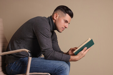 Man with poor posture reading book while sitting on chair against beige background