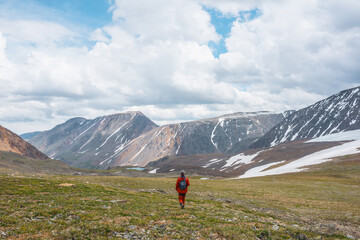 One man in red in sunlit grassy stone field with snow with view to large mountain range in sunlight under cloudy sky. Dramatic mountain landscape with tourist in high mountains at changeable weather.