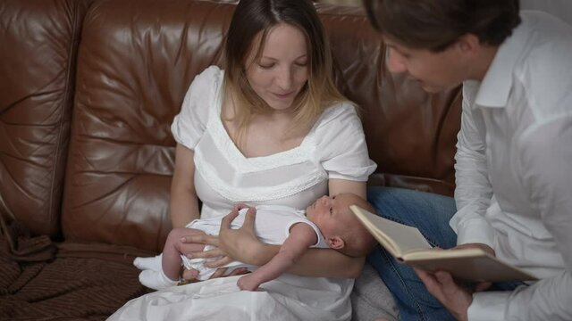 Portrait of Caucasian woman shaking newborn boy in slow motion sitting on couch with man reading fairy tales in book. Smiling mother wife enjoying infant nap time with husband and son