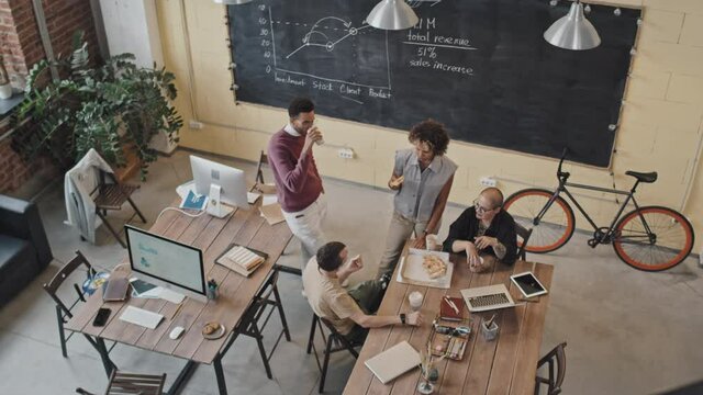 High-angle of four multiethnic female and male colleagues having pizza and coffee, smiling and chatting at table in office in afternoon