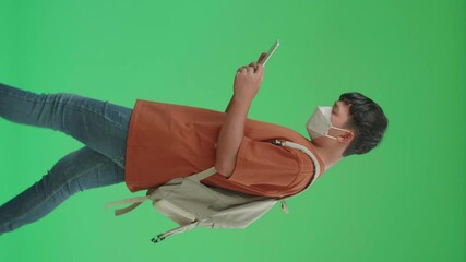 The Side View Of Asian Boy Student Wearing A Mask, Backpack, T-Shirt, And Jeans Using Tablet While Walking To School On Green Screen Chroma Key.

