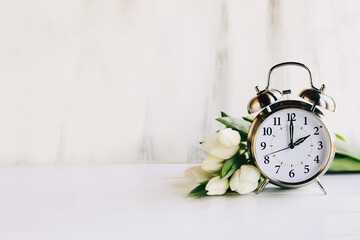 Daylight savings time concept. Set your clocks and to 2 am and spring ahead with this image of an alarm clock with white tulip flowers. Selective focus with blurred foreground and background with copy - Powered by Adobe
