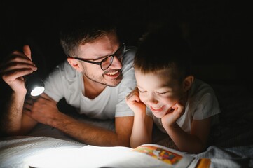 Happy family reading bedtime story under blanket in evening. Father and son spend time together....