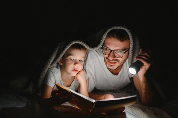 Happy family reading bedtime story under blanket in evening. Father and son spend time together....