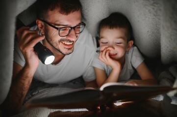 Father and son with flashlight reading book under blanket at home - obrazy, fototapety, plakaty