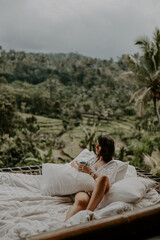 A young travelling woman relaxing in the lounge area of a Bali jungle hotel surrounded by jungle, palm trees and nature