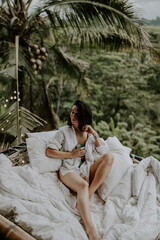 A young travelling woman relaxing in the lounge area of a Bali jungle hotel surrounded by jungle, palm trees and nature