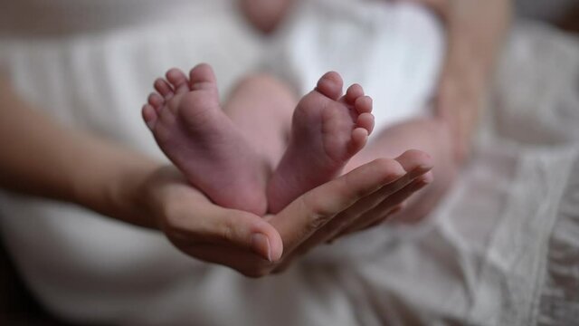 Close-up little Caucasian infant feet in hand of unrecognizable woman indoors. Newborn baby child with caring loving mother at cozy comfortable home. Parenting and love concept. Slow motion