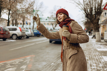Woman with cup of coffee calling taxi and waving hand while standing on roadside in city in winter....