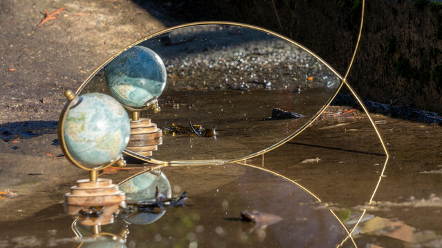 Terrestrial Globe Placed In A Puddle On The Ground, Mirror In The Background, Multiple Reflections