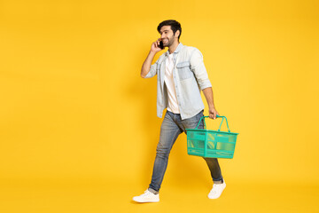 Happy man talk on cell phone and holding shopping basket and mobile phone isolated on yellow studio...