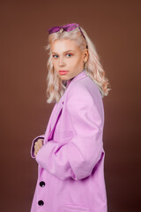Blonde model in a pink jacket with glasses, a jacket with sequins posing in the studio on a brown background. High fashion, 90s 80s. The girl dances and moves. - obrazy, fototapety, plakaty