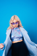 Obraz premium A girl in a blue jacket and purple glasses poses on a blue background and dances. Fashion photo in style 80s 90s party. Blonde girl in a shiny sweater and long earrings