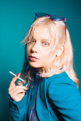 Obraz premium Incredibly fashionable blonde woman in a blue jacket and glasses in the style of 80s 90s poses. On a blue background. Fashionable woman smoking