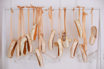 Many hanging ballet shoes on white wall background in studio. New pointe shoes with satin ribbons hanging on rank. Ballet shoes hang on bar in room. Concept of dance, ballet school, ballerinas clothes - obrazy, fototapety, plakaty