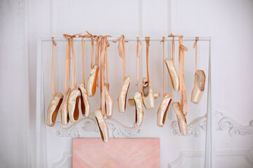 Many hanging ballet shoes on white wall background in studio. New pointe shoes with satin ribbons...