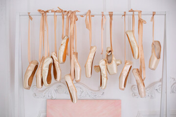 Many hanging ballet shoes on white wall background in studio. New pointe shoes with satin ribbons hanging on rank. Ballet shoes hang on bar in room. Concept of dance, ballet school, ballerinas clothes