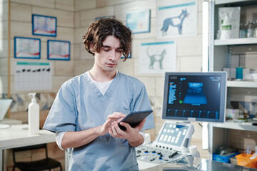 Young vet doctor in uniform typing on digital tablet and working online while standing at his vet...