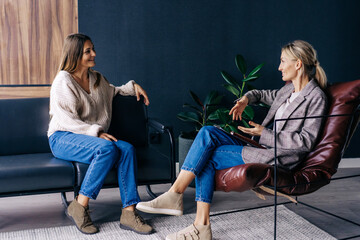 Two modern stylish women are sitting in the office discussing ideas and implementation of plans. A confidential conversation between a woman and a consultant psychologist. - obrazy, fototapety, plakaty