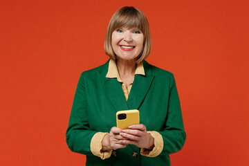 Elderly cheerful happy woman 50s wearing green classic suit hold in hand using mobile cell phone browsing isolated on plain orange color background studio portrait. People business lifestyle concept.
