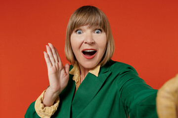 Close up elderly shocked amazed woman 50s wearing green classic suit doing selfie shot pov on mobile phone isolated on plain orange color background studio portrait. People business lifestyle concept