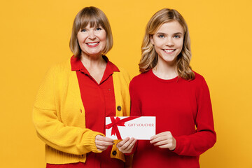 Happy smiling cool woman 50s in red shirt have fun with teenager girl 12-13 years old Grandmother granddaughter hold gift certificate coupon voucher card for store isolated on plain yellow background