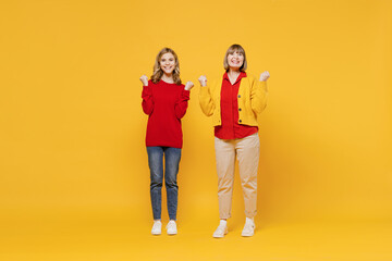 Full body cool woman 50s in red shirt have fun with teenager girl 12-13 years old. Grandmother granddaughter doing winner gesture celebrate clenching fists say yes isolated on plain yellow background