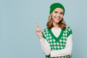 Beautiful excited young brunette girl teen student wear checkered green vest hat point finger aside on workspace area copy space mock up isolated on plain pastel light blue background studio portrait