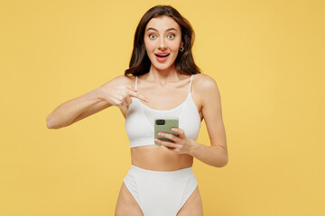 Smiling young brunette woman 20s wearing white underwear with perfect fit body hold use point finger on mobile cell phone chatting browsing internet isolated on plain yellow color background studio.