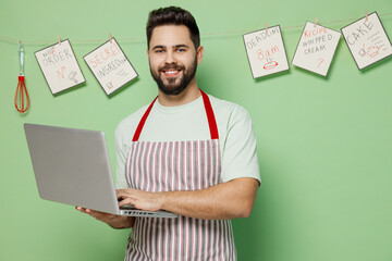 Young smiling satisfied male chef confectioner baker man 20s in striped apron hold use work on laptop pc computer isolated on plain pastel light green background studio portrait. Cooking food concept.