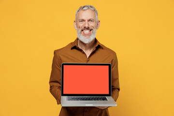 Happy excited elderly gray-haired bearded man 40s years old wears brown shirt hold use work on laptop pc computer with blank screen workspace area isolated on plain yellow background studio portrait.