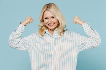 Elderly strong sporty woman 50s wear striped shirt showing biceps muscles on hand demonstrating strength power isolated on plain pastel light blue background studio portrait. People lifestyle concept. - obrazy, fototapety, plakaty