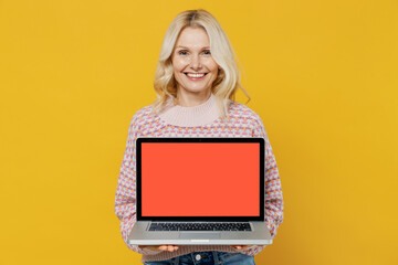 Elderly smiling blonde woman 50s in pink sweater hold use work on laptop pc computer with blank screen workspace area isolated on plain yellow background studio portrait. People lifestyle concept