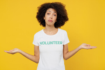 Young woman of African American ethnicity wears white volunteer t-shirt spread hands shrugging shoulders isolated on plain yellow background. Voluntary free work assistance help charity grace concept.
