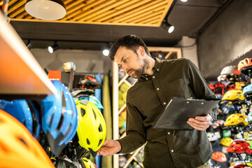 A male bike shop manager makes an inventory of sports helmets in a bike shop. The owner of a sports store with a clipboard in his hands checks the prices of bicycle helmets in the showcase