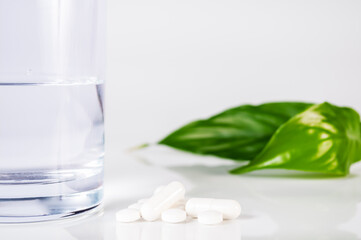 white pills and caplsules, glass of water and green leafs on white background close up