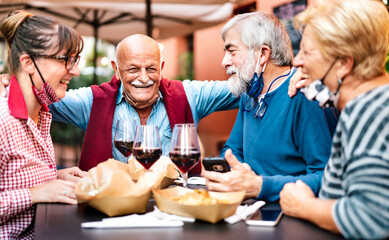 Adult friends drinking wine at restaurant bar wearing open face masks - New normal life style concept with happy mature couples having fun together at home party - Vivid filter with focus on left man