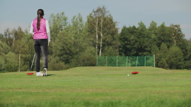 Girl in sunglasses and a pink vest hits a ball with a club while playing golf on a green golf course. A woman chooses a golf ball for long distance and looks into distance and excitedly pats her leg.