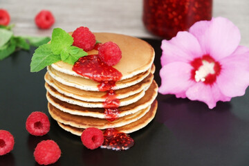 Pancakes with fresh raspberries, mint and jam on a black table with lonicera