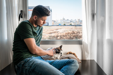 young man sitting by the window strokes the head of a black  and white cat