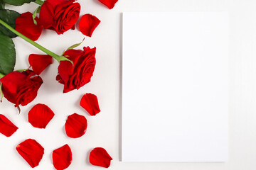 Greeting card mockup with red roses and petals on white wooden table top view. Template with copy space for design for Valentine's Day, mother's day, wedding, flat lay.