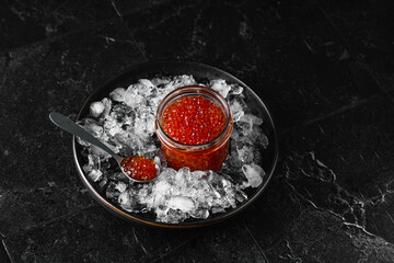 Red salmon caviar in a glass jar on crushed ice. Black background. Dark photo