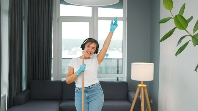 Woman in headphones cleans the house and have fun singing with a broom like a star at a concert. Slow motion