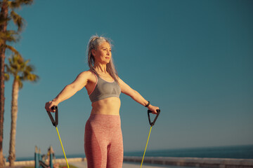 Portrait of mature female exercising with fitness resistance bands standing on the sports site on seashore in the evening, watching the sunset