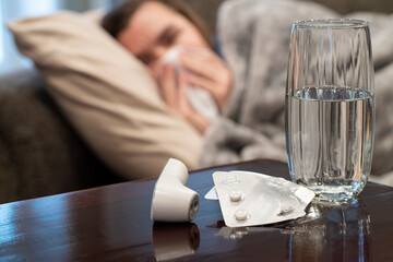 Cold medication with water and thermometer and person with covid cold or flu blowing their nose in background