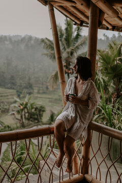 Young female traveller is enjoying a cup of coffee on a terrace in her boutique hotel in Bali, Indonesia. Surrounded by lush green jungle, rice fields, palm trees 