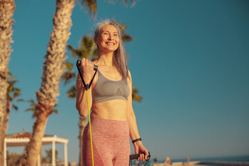 Happy Asian lady exercising using rubber bands bending her arm at elbow, standing against the background of palm trees, smiling