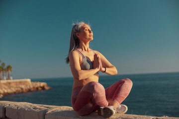 Fototapeta na wymiar Middle-aged sporty woman in sportswear sitting near the seashore in lotus position and training on sunny day