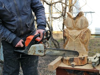 Wood artist with a chainsaw carves a solid wood owl statue, work on processing wood material with a chainsaw in a rural garden, creating interior and design items from solid wood with a hand tool