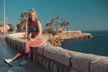 Fototapeta na wymiar Smiling sporty lady in sports top and pink leggings sitting by the seashore and holding bottle of water